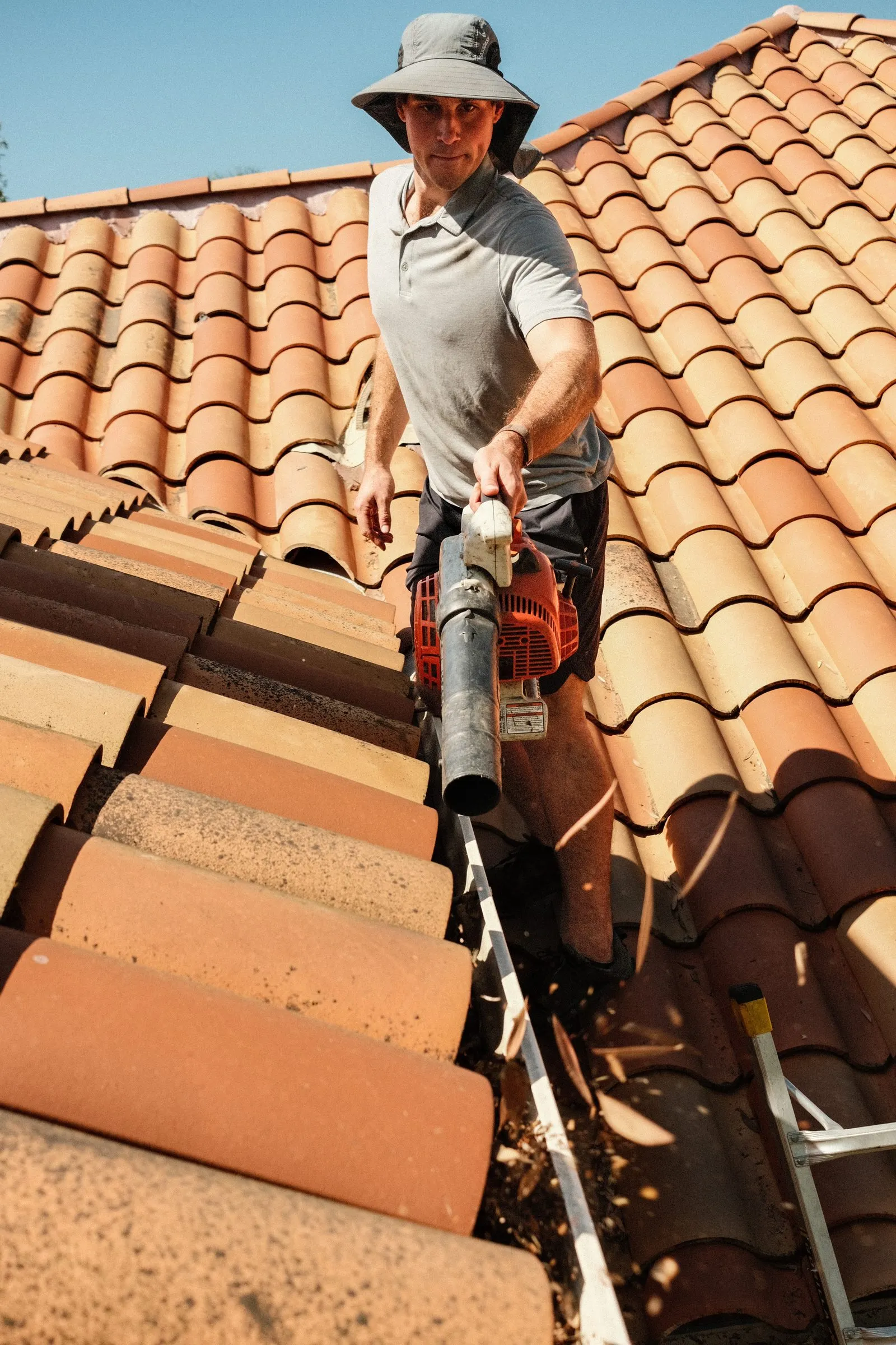 Elliot on a tile roof facing the camera, blower in the gutter, debris flying