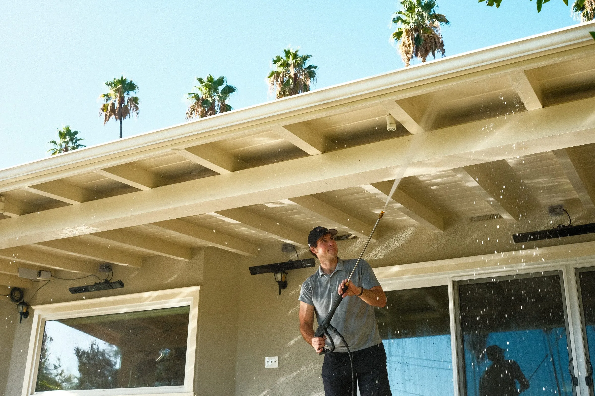 Elliot pressure washing under a patio cover, spray in the air, a row of palm trees above