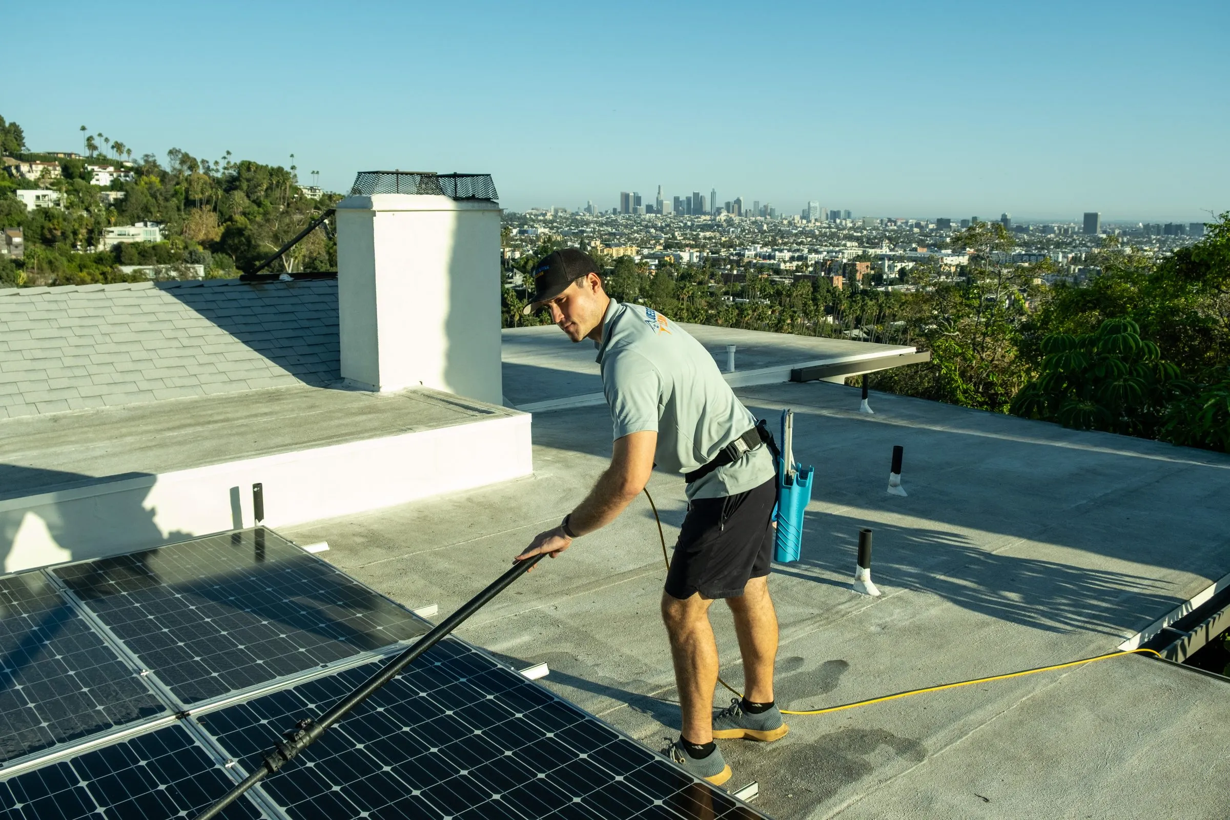 Crew member brushing rooftop solar panels with a water-fed pole on a flat roof, the city skyline in the distance
