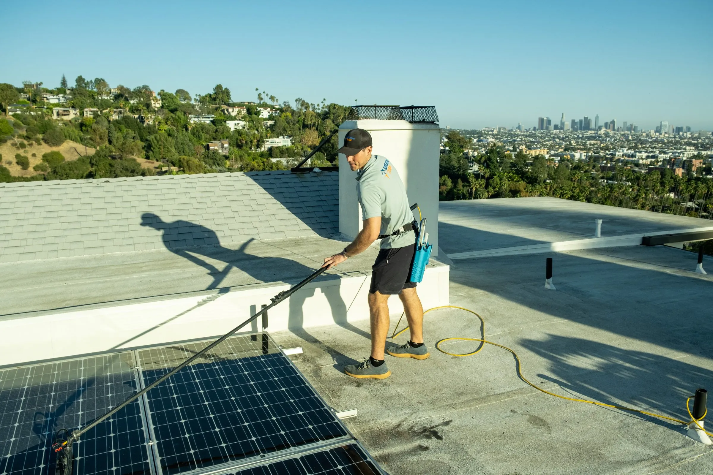 Crew member guiding a water-fed pole onto rooftop solar panels in low sun, a long shadow across the roof, the city skyline on the horizon