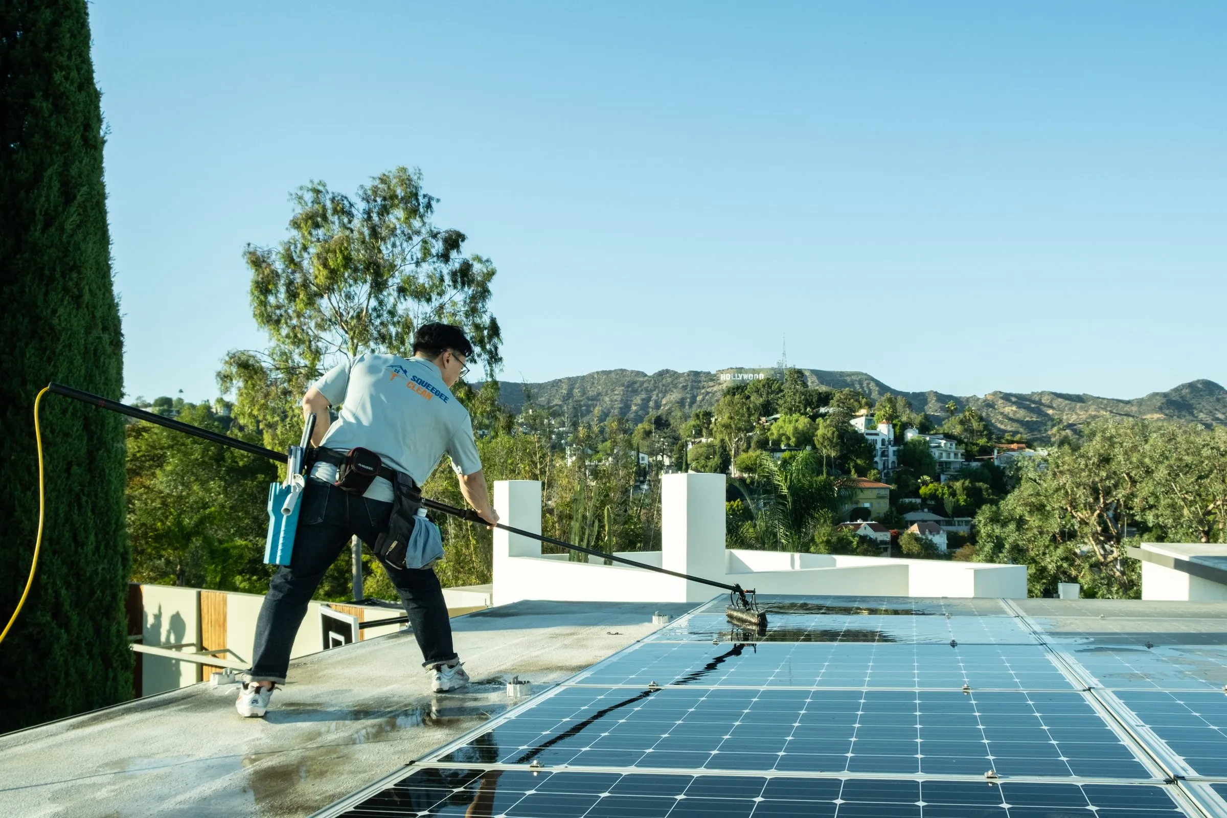 Water-fed pole stretched across a row of rooftop solar panels, hills behind