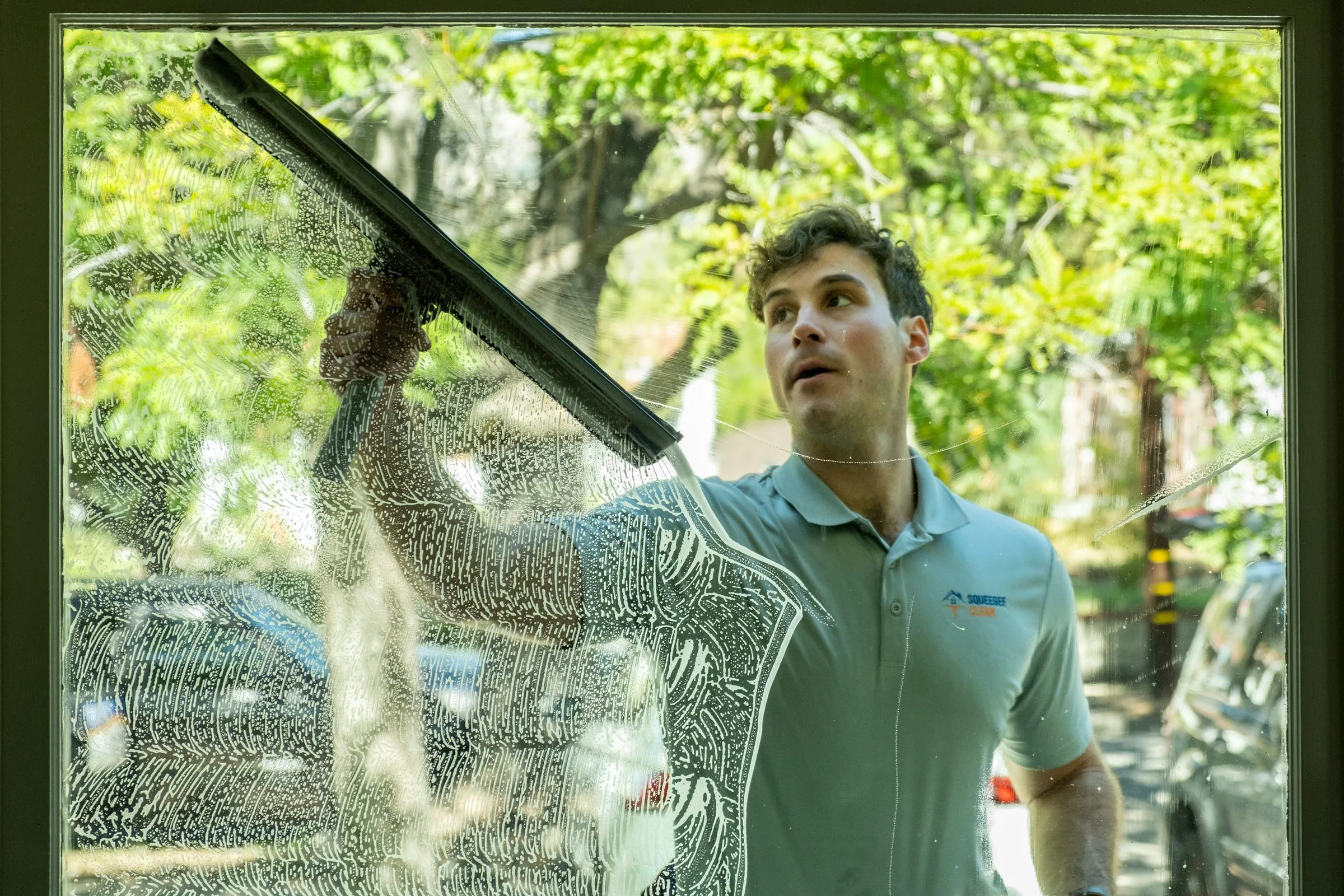 Elliott squeegeeing a soapy window seen from inside, trees reflected in the clean half