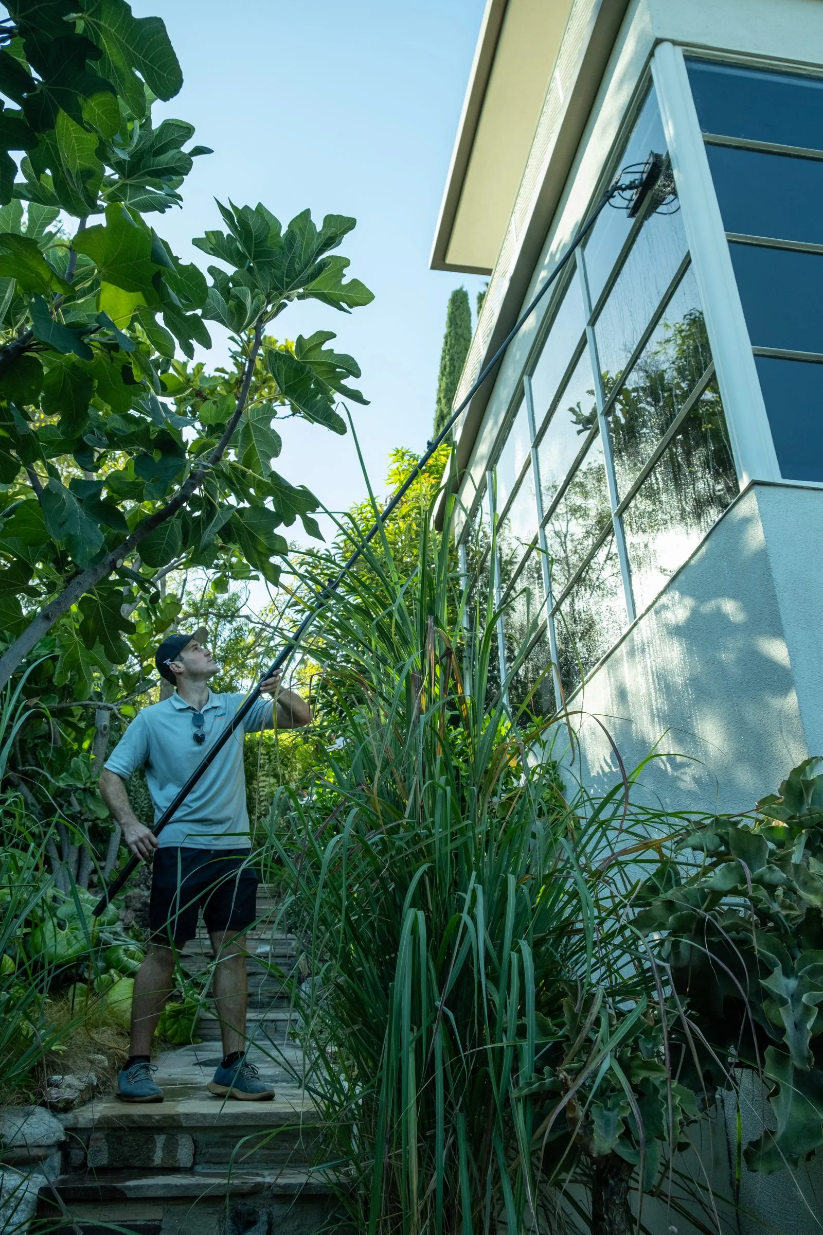 Wes working a water-fed pole up a modern house with floor-to-ceiling glass, tall grasses in the garden